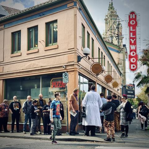A human line forms around a building, with Hollywood Theatre and its vertical sign in the distance on a sunny day.