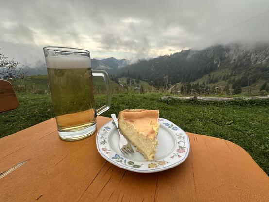 Das Bild zeigt eine idyllische Szene im Freien, wahrscheinlich in einer Bergregion. Auf einem orangefarbenen Holztisch steht ein großes Glas Bier mit Schaumkronen, daneben ein Stück Käsekuchen auf einem weißen Teller mit blühendem Rand. Der Teller ist mit einer Gabel versehen. Im Hintergrund erstreckt sich eine malerische Landschaft mit grünen Hügeln und bewaldeten Bergen, die von Nebel umhüllt sind. Der Himmel ist bedeckt mit Wolken, was auf einen bewölkten Tag hinweist. Die Szene vermittelt ein Gefühl von Ruhe und Entspannung, ideal für einen entspannten Moment im Freien.