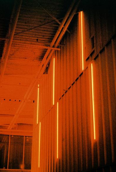 Black and red image of an industrial space. 6 strips of light illuminate the interior of the corrugated steel structure from the wall on the left of the image.