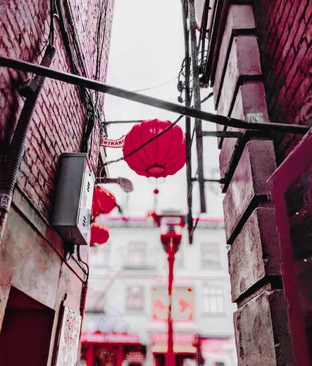 Looking up an alleyway between brick walls, a vibrant red Chinese lantern hangs overhead, with blurred background of Chinatown architecture.