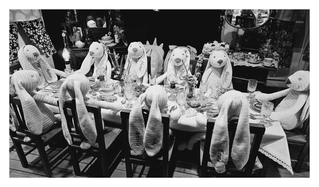 A black and white photo, oriented horizontally, shows a dinner table set for a tea party. Seated around the table are several plush rabbits, each with a name embroidered on its ears, such as "Markus," "Ofelia," "Theodor," and "Andreas." The rabbits are wearing pajamas or casual clothing. The table is covered with a white tablecloth and set with plates, glasses, and teacups. In the background, there's a buffet and a large mirror.