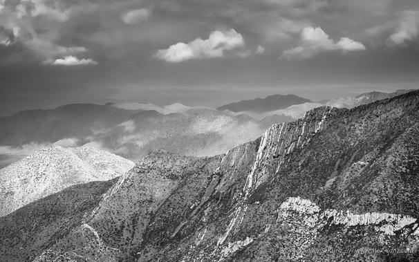A black & white landscape photo made from an elevated viewpoint, looking across barren ridges of a desert landscape under monsoon clouds. Light breaks through the clouds here and there, painting light and shadow onto the landscape.