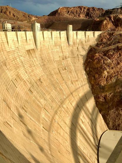 A large concrete dam is shown, with a textured surface reflecting sunlight. The surrounding landscape features rocky terrain, and power lines are visible in the background. The dam's structure creates a significant shadow on one side, emphasizing its imposing height.