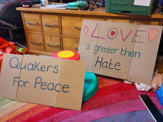Two cardboard, handwritten signs propped up against a console table in a room with a rainbow carpet. The nearest sign reads "Quakers For Peace". The other reads "LOVE is greater than Hate" and has several hearts drawn around it.