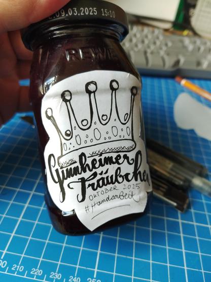 A jar of dark red grape jam with a handmade calligraphy sign saying "Ginnheimer Träubchen" and a crown. All on a blue work surface with some pens and tools.