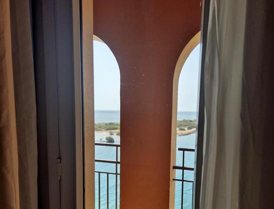 Oben abgerundete Fenster mit halb zugezogenen Gardinen.
Durch die Fenster sieht man auf Meer und bewachsene Landbereiche.