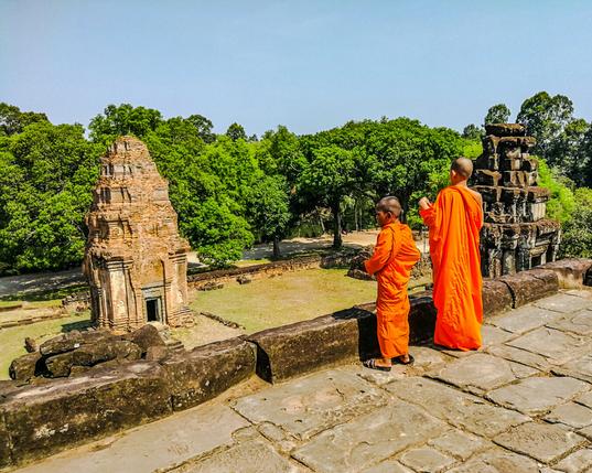 Photo shows two young novice Buddhist monks in deep orange robes on top of a temple structure looking out to a landscape covered in deep, thick green forest. In front of the then, they can look down on a temple tower in early Khmer imperial style in good condition. To their right, an equal tall temple tower on the same level as the first are in clearly less renovated. Darker areas, worn down details and more hidden reliefs gives the impression of how this must have looked when Bakong temple was rediscovered. It is a sunny morning, with clear blue sky. Cambodia is as safe, comfortable and kind as ever. Angkor and Siem Reap welcomes visitors. Enjoy your visit to the Kingdom of Wonder.