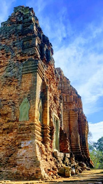 Photo shows the side of two brick temple towers in early Khmer style. The one closest to the camera is restored with wall arts and stone details of high quality, but still with some darker details and stone near the top. The dancing apsaras reliefs are visible, but the one closest is mostly a green-grey outline. Its neighbour tower is more completely renovated, and the red bricks on the top is clean red. It is early on a sunny day with slightly overcast blue sky. The morning sun gives the towers a warm yellow-red glow. In the background there are some distant green treetops contrasting the reddish towers. Cambodia is as safe, comfortable and kind as ever. Angkor and Siem Reap welcomes visitors. Enjoy your visit to the Kingdom of Wonder.