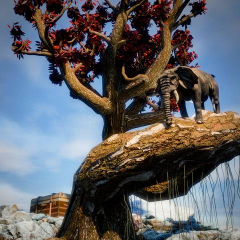 The image shows a surreal sculptural scene photographed from below against a blue sky. An elephant stands to the right atop a large, twisted tree trunk with a textured, mottled surface in browns and golds with patches of white. Behind and around the elephant, gnarled branches with golden-brown bark extend upward and outward in dramatic, twisting formations, supporting dense clusters of dark leaves with striking red-orange flowers or blooms that create vivid contrast against the blue sky. Long green grass-like strands hang down from beneath the trunk like natural fringe or roots. At the base of the composition, a brown travel trunk with leather straps sits among scattered white rocks or stones. The entire scene has a fantastical, dreamlike quality with its unusual scale, twisted organic forms, and the whimsical placement of the elephant high above the ground on the contorted tree trunk.