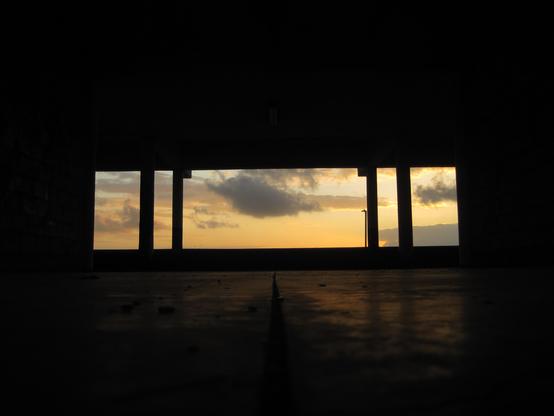 Fotografía tomada desde un lugar oscuro que enmarca la luz del amanecer. En primer plano, el suelo y el interior del espacio, posiblemente un porche o una estructura con techo, están en sombra, creando un fuerte contraste con el exterior brillante.

La toma está hecha desde un ángulo bajo, con una línea oscura que parece ser una junta o grieta que divide el suelo en el centro y se dirige hacia el horizonte.

El amanecer es la estrella de la imagen, visto a través de una abertura rectangular, dividida por dos grandes postes o columnas, y otros más delgados a la derecha. El cielo muestra una mezcla de colores cálidos: tonos dorados y anaranjados cerca del horizonte, que se mezclan con amarillos pálidos y toques de azul y gris en las nubes. Una nube más grande se asienta un poco más arriba, rompiendo la uniformidad del cielo.

Hacia la derecha, se distingue la silueta oscura de un poste de luz o farola, acentuando la atmósfera de transición entre la noche y el día. La composición es dramática, con los elementos oscuros del interior funcionando como un marco natural para la explosión de color del alba.