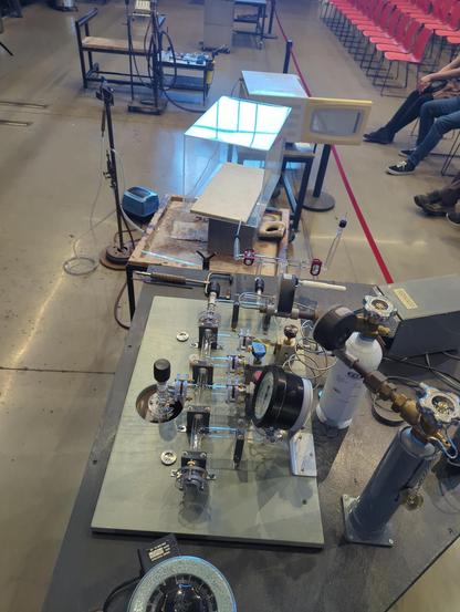 Chrysler's fancy new manifold on a cart with a clear acrylic box next to it and a microwave behind that. All behind the red tape in front of a public audence on the hot glass studio floor