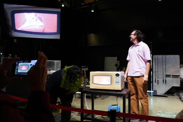 me looking at the big screen and smiling as the evacuated and backfilled neon glass lights up inside of the microwave and Abbey checks on it. An audience member snaps a photo on their phone.