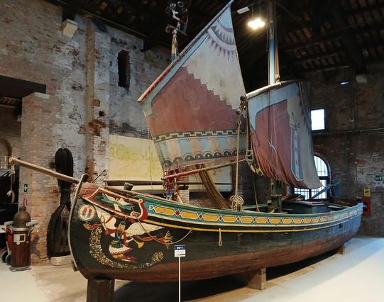 A wooden two-masted fishing schooner in Venice's naval history museum, with a black hull topped by a patterned yellow stripe and an angel blowing a horn pained on the bow. Her lug sails are also gaily decorated in red, blue and yellow patterns.