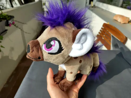 A small hyena plushie held by a hand, well my hand no less! They got an awesome purple punk mane and purple tail tuft.