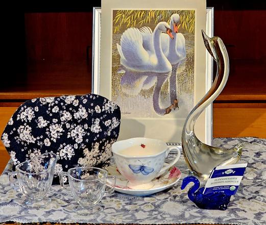 Tea tableau backed by a print of Robert Gillmor's painting of a pair of Mute Swans floating on a pond. There is also a tall clear Murano glass swan sculpture, a small Mosser cobalt glass swan salt cellar, an Art Deco clear glass cream and sugar from the 1920s, a Lenox Butterfly Meadow pedestal cup, a navy blue chintz tea cozy, and Elderberry herbal tea.