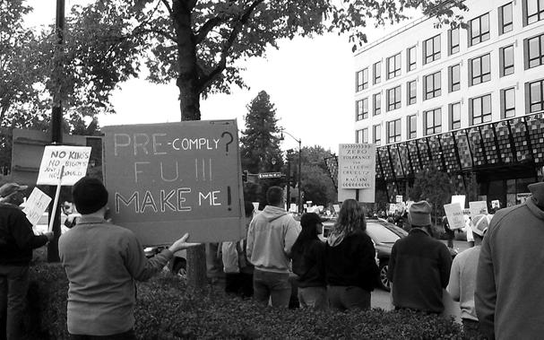 Black and white photo of protestor holding a sign "Pre-comply? FU!! Make Me!" amongst other fellow protesters holding various signs.