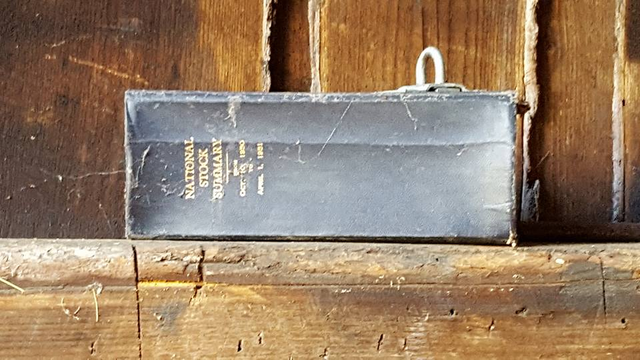 A thick black book rests on a beam in an old barn. It has a leather cover that's tattered  along the opening cover. The book is called National Stock Summary and the date is October 1950 to April 1951. Behind the book are the vertical boards of a barn wall. There's wisps of spider webs to the right of the book.