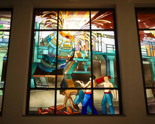 Vitrail représentant le travail des hauts fourneaux, avec des ouvriers travaillant dans une usine. La Haut fourneau illumine le vitrail Vitrail représentant le travail des hauts fourneaux, avec des ouvriers travaillant dans une usine. La Haut fourneau illumine le vitrail