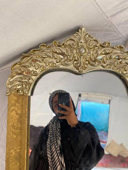 Selfie of Naeema in front of a mirror in the tent. Her face is partially hidden behind the phone. She's wearing a keffieh. Selfie of Naeema in front of a mirror in the tent. Her face is partially hidden behind the phone. She's wearing a keffieh.