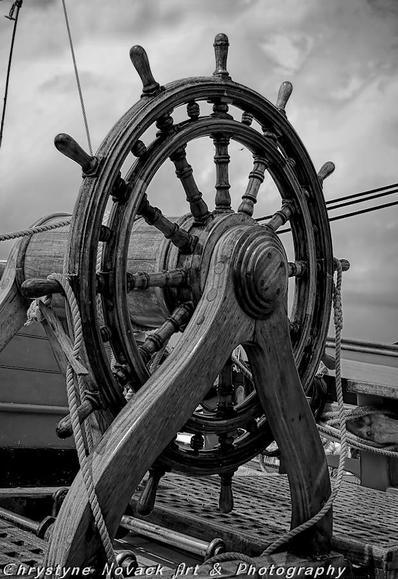 This black and white photo captures all of the details of the tall ship HMS Bounty steering wheel inviting the viewer to take the helm. The black and white captures the detail of the grain in the from the handles to the felloe to the spokes. And by sheer luck a seabird in the distance can be seen flying between the two upper spokes of the wheel. This black and white photo captures all of the details of the tall ship HMS Bounty steering wheel inviting the viewer to take the helm. The black and white captures the detail of the grain in the from the handles to the felloe to the spokes. And by sheer luck a seabird in the distance can be seen flying between the two upper spokes of the wheel.