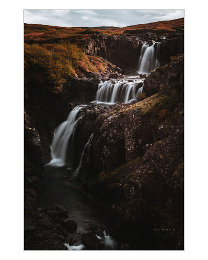 A soft, misty waterfall descends over dark rocks surrounded by moss and autumn light. The slow flow of water creates a sense of stillness and time suspended. The quote “I am rooted, but I flow.” by Virginia Woolf appears discreetly over the image captured by Gus Martinié