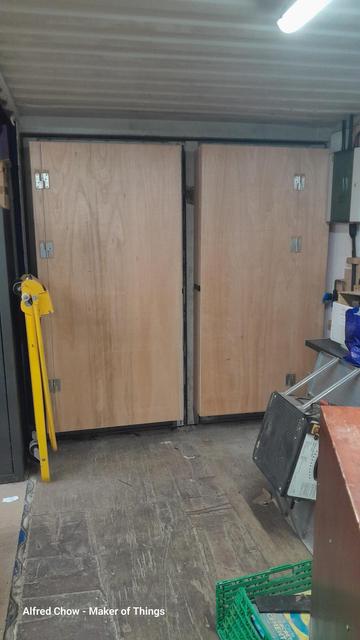 The corner of a large workshop built from two shipping containers. Seen from inside, the container doors look like they have been clad in plywood. But hinges on the plywood hint at something else, something hidden.