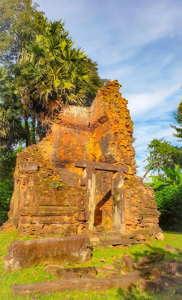 Photo shows the remains of a single tower temple built in old Khmer Hindu style closely resemble the older Chenla style. The material is red laterite stone, and in the sunrise, it makes the old temple glow warm yellow-red. In the background there are palm trees and some other forest trees, and the sky is blue with some light clouds. The temple is nearly squared but has lost its top/roof. On two sides the old walls reach nearly up to original heights. But the rest of the walls has fallen, leaving a human tall perimeter wall and the temple door gate is the tallest remaining structure. Some stones from the temple are laying on the ground around. Cambodia is as safe, comfortable and kind as ever. Angkor and Siem Reap welcomes visitors. Enjoy your visit to the Kingdom of Wonder.