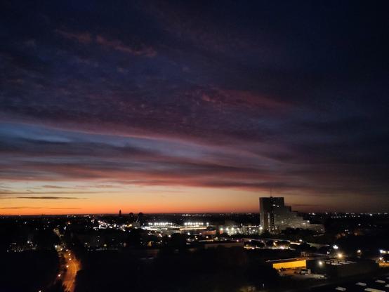 levé de soleil pastel, avec du bleu et du orange, plus les nuages qui forment un pattern doux, et la ville sombre aux petites lumière orange et jaune. un grand batiment se découpe a droite en forme de marchés d'escalier, mais vu d'ici il a l'air tout petit