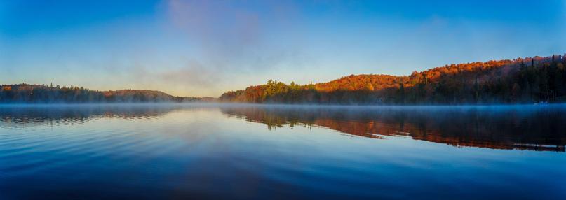 A serene lake at dawn, reflecting blue skies and distant trees, with a light mist hovering over the water's surface.