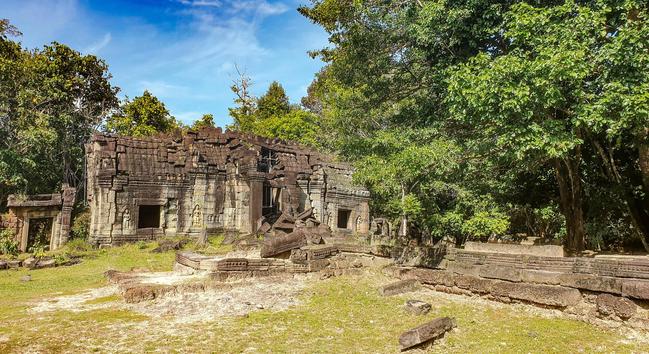 Photo shows the entrance gate of the Great Citadel, Banteay Thom, a massive but low entrance structure with five gate door holes. The structure is made of brickwork and laterite stone. The colours are light grey, light brown, dark brown and some red stones. In front there is a stone made plateau/terrace. At the wall of the entrance there are stone carvings of dancing Apsara figures, text and some art patterns. The entrance is framed by green trees and turf. A lightly clouded deep blue sky tells of late wet season. Cambodia is as safe, comfortable and kind as ever. Angkor and Siem Reap welcomes visitors. Enjoy your visit to the Kingdom of Wonder.