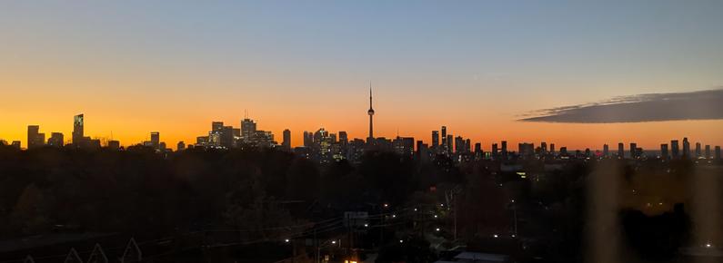 A sunrise over Toronto’s skyline, looking south from midtown. The city buildings, with the CN Tower in the middle, are in almost complete darkness.  There is a band of orange sky above them, transitioning to blue sky higher up.