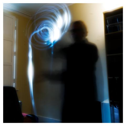 The image depicts a dark silhouette of a person in a brightly lit room. The silhouette appears to be standing against a pale yellow wall, with a dark cabinet or bookshelf visible to the right. Emanating from the person's head are bright, swirling blue light trails that create a dynamic, almost ethereal effect. The light trails are blurred due to motion, suggesting movement or a long exposure photograph. Parts of a chair are visible in the bottom left corner of the frame.