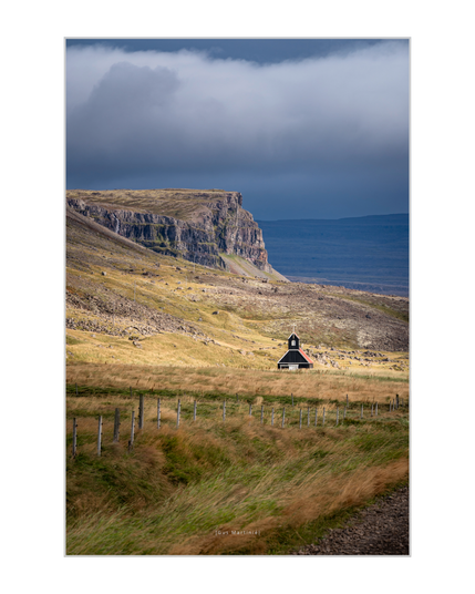A small black wooden church with a red roof stands in sunlight alone in a golden Icelandic field beneath a stormy sky. Massive cliffs rise behind it, and wind bends the grass in waves — a quiet image of solitude and shelter. By Gus Martinie