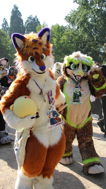 Photo of Fursuiters Fox Flummox carrying a very roumd plush chicken and dragging Gustl by the Paw. Gustl is waving. Other Fursuiters behind them of which none is recognizable. Park trees in the background. Sunlight from the back makes the fur outline glow very bright