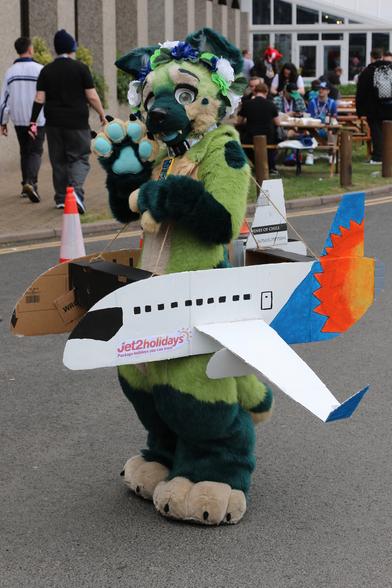 Photo of a green dog fursuiter wearing a cardboard cutout in the crude shape of an aircraft, painted in the livery of a package holiday operator.