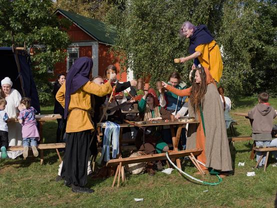 This is a photo of an open-air live performance at medieval-style fair. The image depicts a group of people dressed in historical or medieval-style clothing gathered outdoors in a grassy area. They appear to be participating in a social or communal activity, possibly a festival or reenactment. The individuals are standing and sitting around wooden tables, engaging in what seems to be a toast or a ceremonial gesture, as they hold up cups or mugs. Some are wearing head coverings, and the attire includes tunics, cloaks, and hats, suggesting a historical or fantasy theme. The background features a red brick building with a green roof, surrounded by trees and greenery. The overall atmosphere suggests a historical or cultural event.