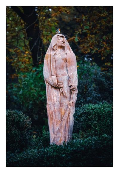 This photo is of a weathered, stone statue of a robed woman standing in the middle amidst greenery. The statue has aerene expression, holds some of the fabric of the robe in her right hand and the cloth from the robe drapes over her head. A small red robin is perched on her head. The background has dense, dark green foliage with little red/orange tints of autumn colors.