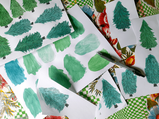 Sheets and paper scraps with illustrations of conifer trees on them lying on a table. In the middle is a sheet with just green blobs painted on it in different colours. On the other papers details have been drawn on the green blobs with a black pen to create trees. To the right is lying a black pen.