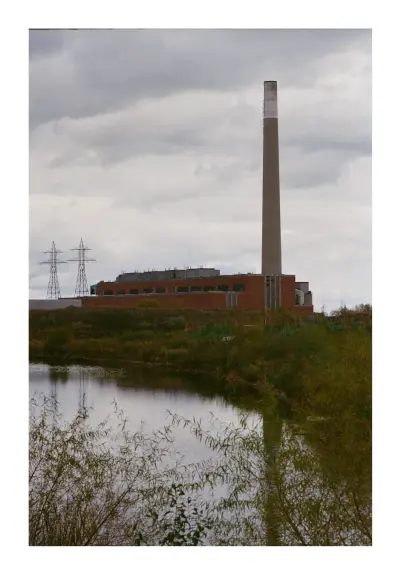 A colour photograph of the Hern generating station, a decommissioned coal fired power plant, situated in the Portlands of Toronto viewed from Biidaasige Park, a new urban park in the reclaimed industrial lands and re-naturalized mouth of the Don river. 

The foreground is the new naturalized wetlands in the background is the turbine house and smokestack and electricity transmission towers.