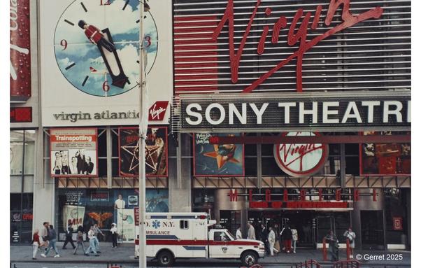 Street scene in front of a Virgin-branded Sony Theatre. Large Virgin Atlantic clock and movie posters visible. Busy sidewalk with pedestrians and NY ambulance.