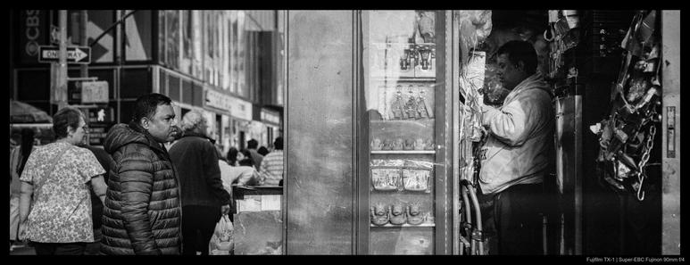 A pedestrian stands stopped amidst a moving crowd to look at the front of a convenience kiosk.  The photo is angled such that both the passerby and the shopkeeper are visible; the latter is standing in the darkened interior of the stall.