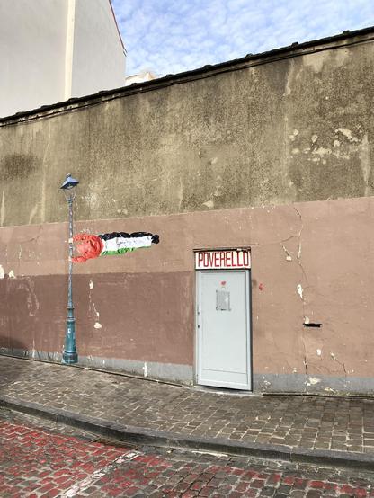 Mur avec porte d'accès au "Poverello" (centre d'accueil pour sans-abris) avec à côté un graffiti mural montrant un gymnaste vêtu aux couleurs de la Palestine se tenant  à un réverbère, devenant ainsi lui-même une sorte de drapeau.