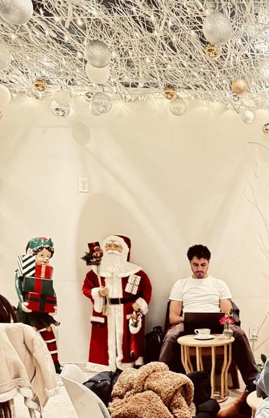 Color photo taken inside of a coffee shop where many people go to do work on their laptop computers. This coffee shop has gone to town on the Christmas decorations. The ceiling is covered in white vines with little white sparkly lights and silver and gold ornament balls. Below this, sitting against a white wall is a man looking down at his laptop doing his work with a cup of coffee on the table in front of him. Right next to him on the left is a 4 foot tall Santa statue that is the same height as the man sitting down. On the other side of the Santa is an elf statue the same size. Some coats thrown on chairs can be seen at the bottom of the photo