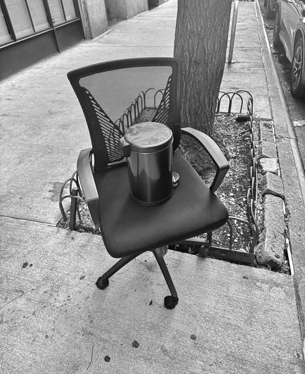 Black and white photo of a black office chair with a mesh high back and arms. Sitting on the chair is a small black lidded trashcan with dust all over it. The chair is sitting with one wheel in the square piece of dirt surrounding a tree on a city street. More sidewalk in the bottom of some windows can be seen in the background