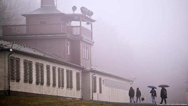 People holding umbrellas walk in the rain up the entrance to Buchenwald concentration camp.
