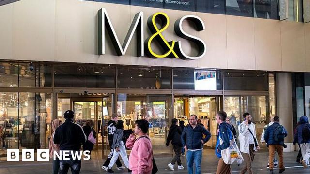 A crowd of people walk outside of a large M&S store with an M&S sign hanging overhead