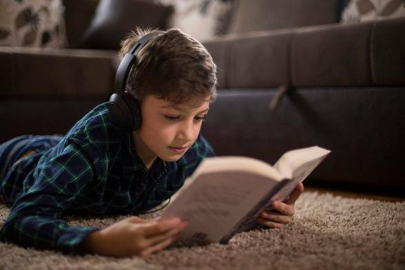 A child with headephones on is lying on a rug and reading a book