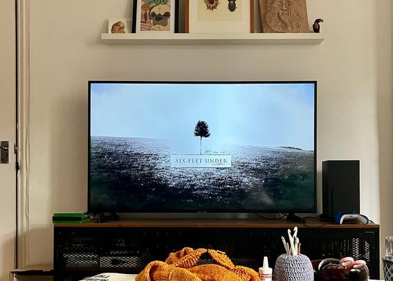 TV on a table against a wall, showing the title card for Six Feet Under. Several items on a coffee table in the foreground, a small shelf above the TV with various art pieces.
