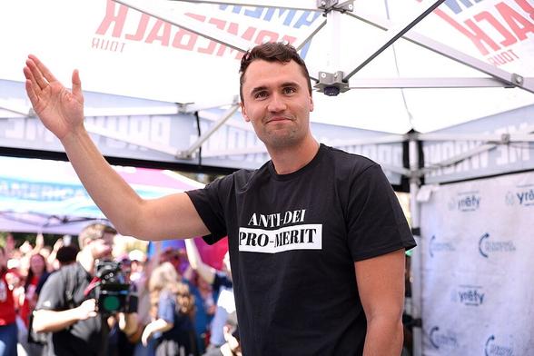 Charlie Kirk stands under an outdoor tent waving. He wears a black and white shirt with the text “ANTI-DEI PRO-MERIT”.