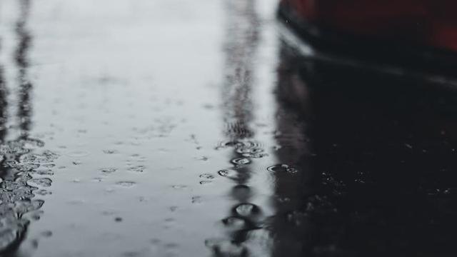 Close-up of rainwater droplets scattered on a smooth, dark surface, creating a reflective, blurred effect with indistinct vertical shapes in the background.